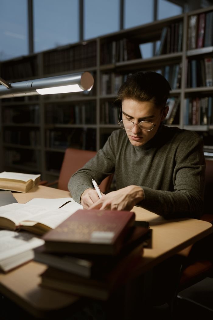 services-01 A young man in a library studying with books and taking notes under a desk lamp.